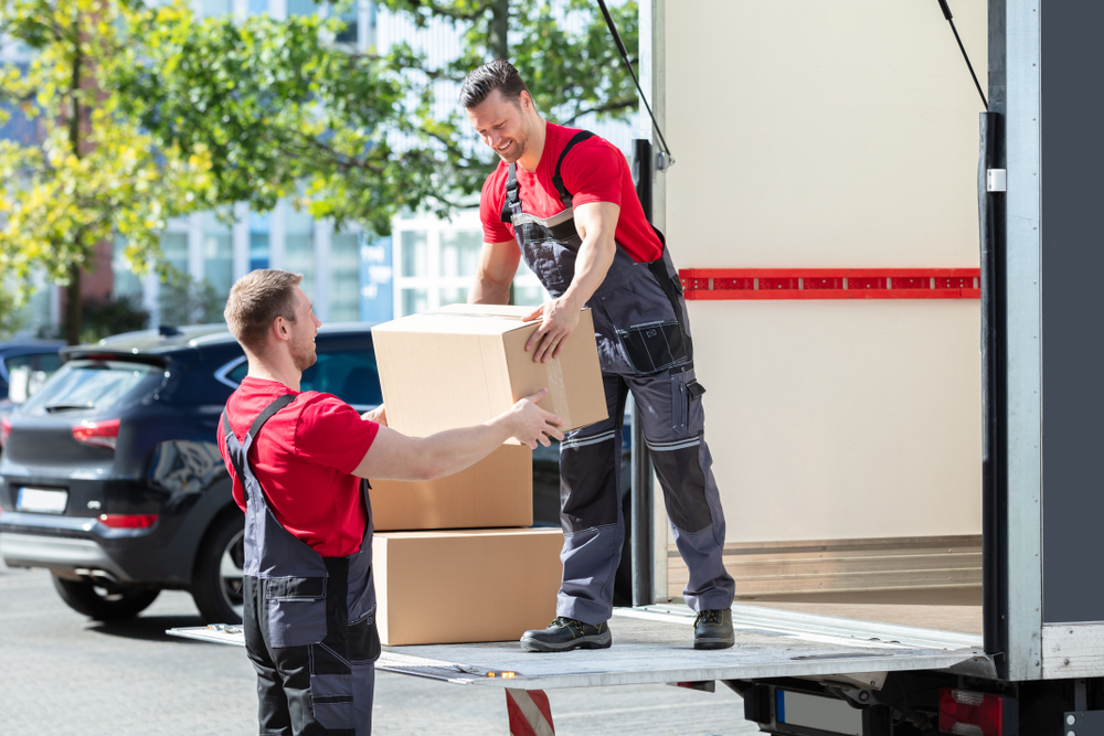 Two movers in red shirts and overalls, serving Lake & Orange County, FL, are unloading cardboard boxes from a truck. One stands on the lift gate passing a box to the other on the ground, with cars and trees visible in the background.