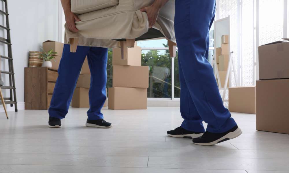 Two people dressed in blue pants and black sneakers, likely movers Lake & Orange County, carry a beige armchair in a room filled with cardboard moving boxes, with light streaming in through a window.