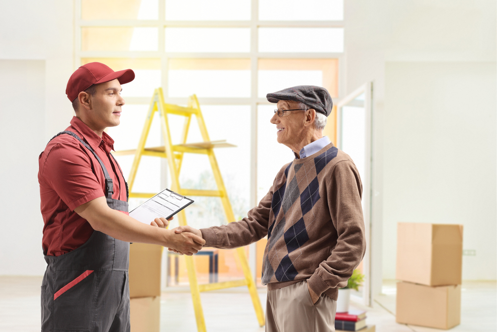 A delivery worker in uniform shakes hands with an older man wearing glasses and a cap, inside a room with moving boxes, a clipboard, and a yellow ladder—trusted movers Lake & Orange County, FL ensuring a smooth move.