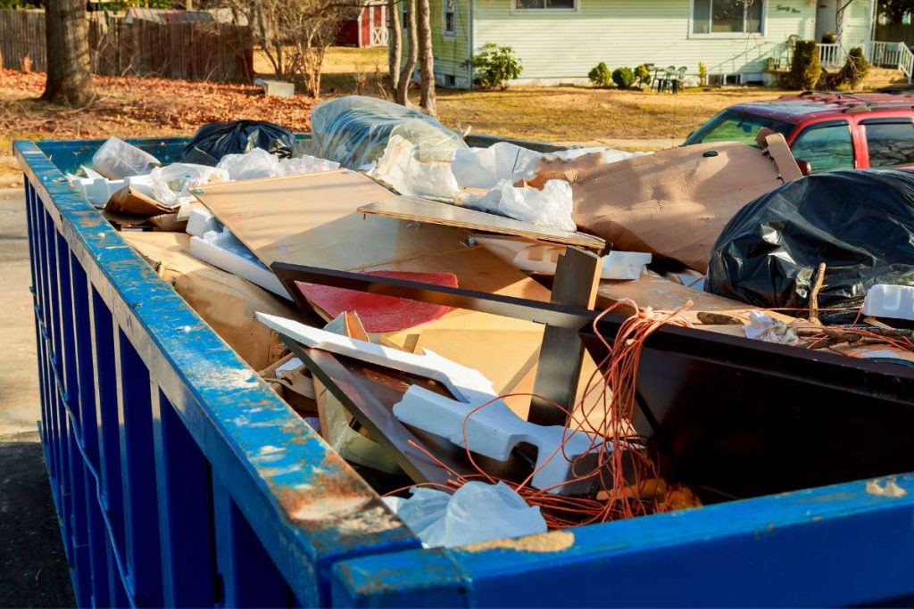 A large blue dumpster filled with debris, including wood, plastic bags, and furniture pieces, sits outdoors near a residential area in FL. Movers Lake & Orange County services are nearby, with houses and parked cars visible in the background.